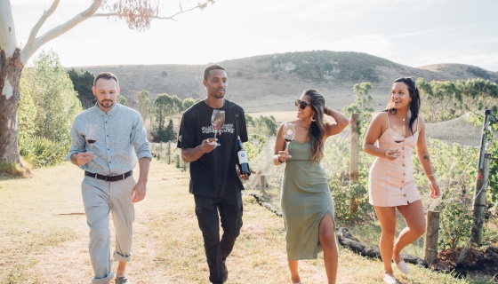 A group of friends holding glasses and walking through a vineyard on Waiheke Island, Auckland